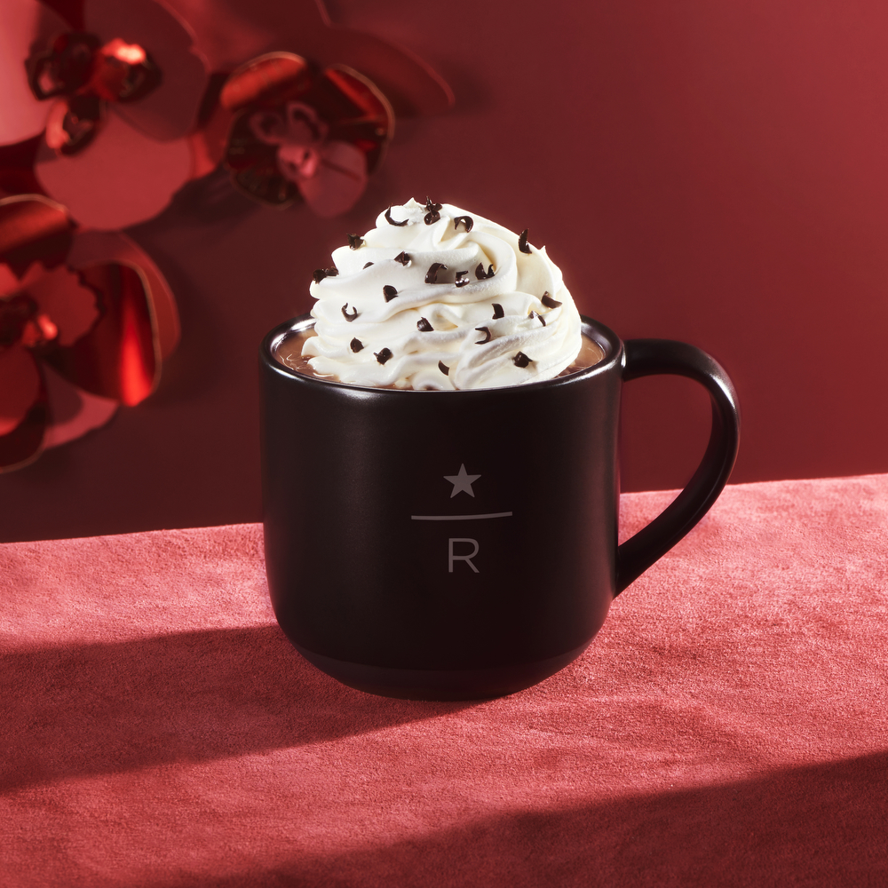 Starbucks Reserve mug containing beverage topped with whipped cream and chocolate shavings, on a red tablecloth with red backdrop.