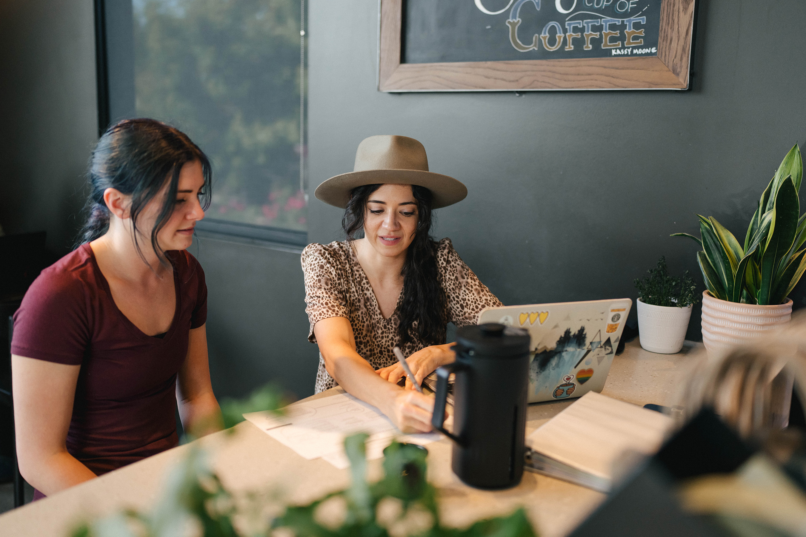 Woman with long hair and hat holding a pen, showing another person something on paper.