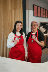 A gif of two Starbucks barista wearing red apron and holding a red reusable cup and cheering together