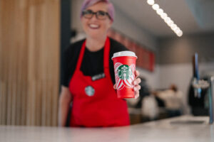 A Starbucks barista in a red apron handing over a red reusable cup