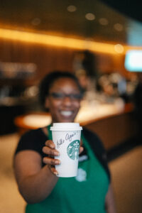 A Starbucks barista holding a Starbucks cup with writing on it that says Hello Again!