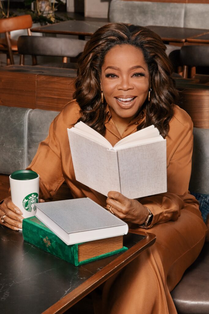 Oprah sitting in a Starbucks store holding a book and a Starbucks mug