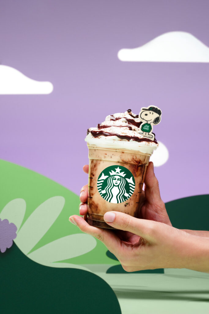 A hand holding a cup of a frappuccino topped with whipped cream for Starbucks and Peanuts collaboration