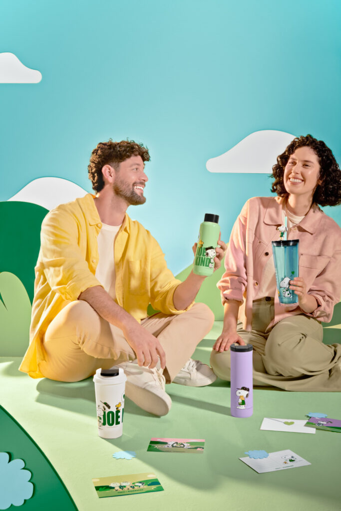 A male and a female sitting down smiling with cups in their hands for Starbucks and Peanuts collaboration