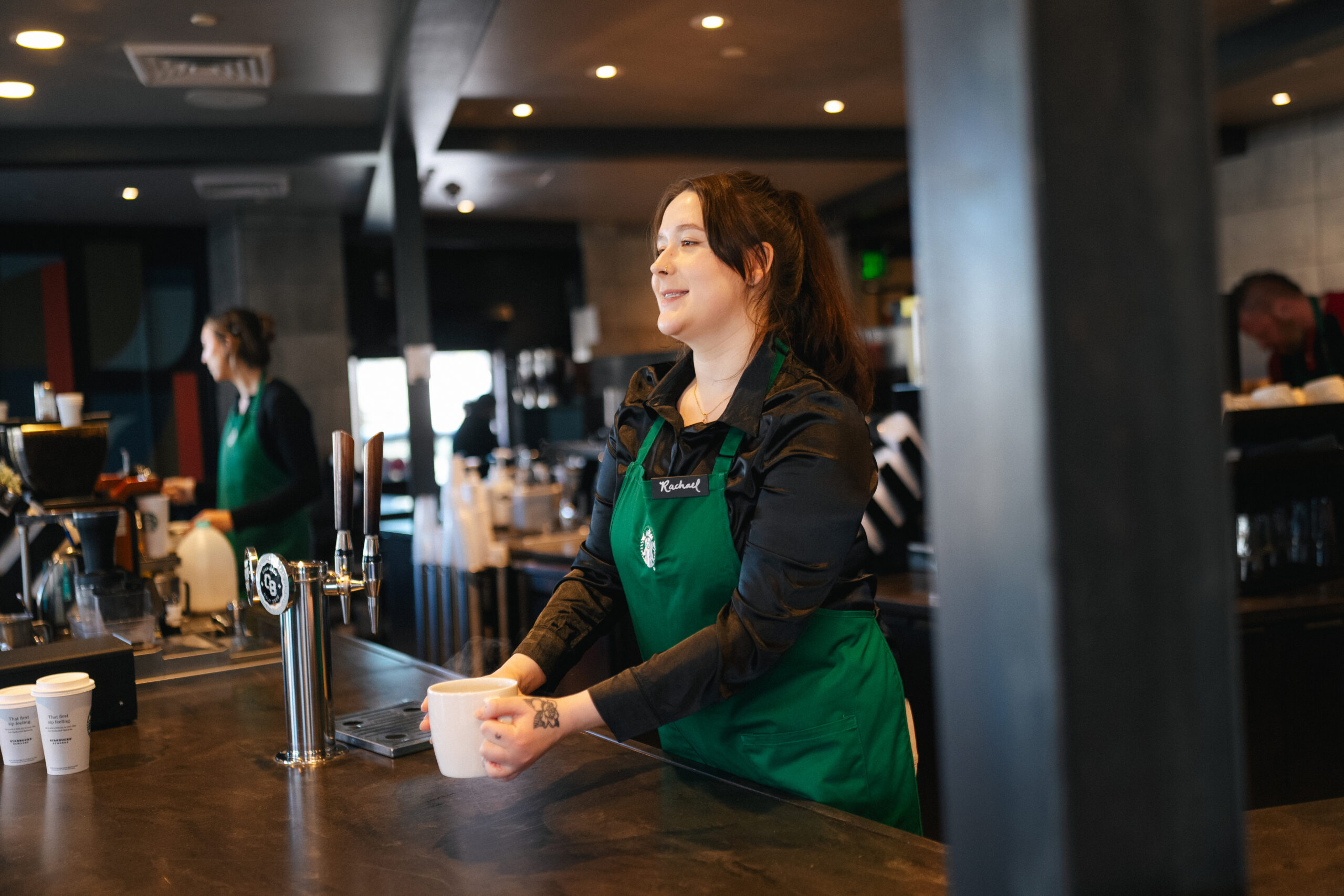 A barista in a green apron stands behind a coffee counter, holding a steaming white cup. She has long brown hair tied back and is smiling, appearing engaged in her work. The background features a modern café setting with various coffee equipment and another barista visible in the distance. The atmosphere is warm and inviting, with soft lighting illuminating the space.