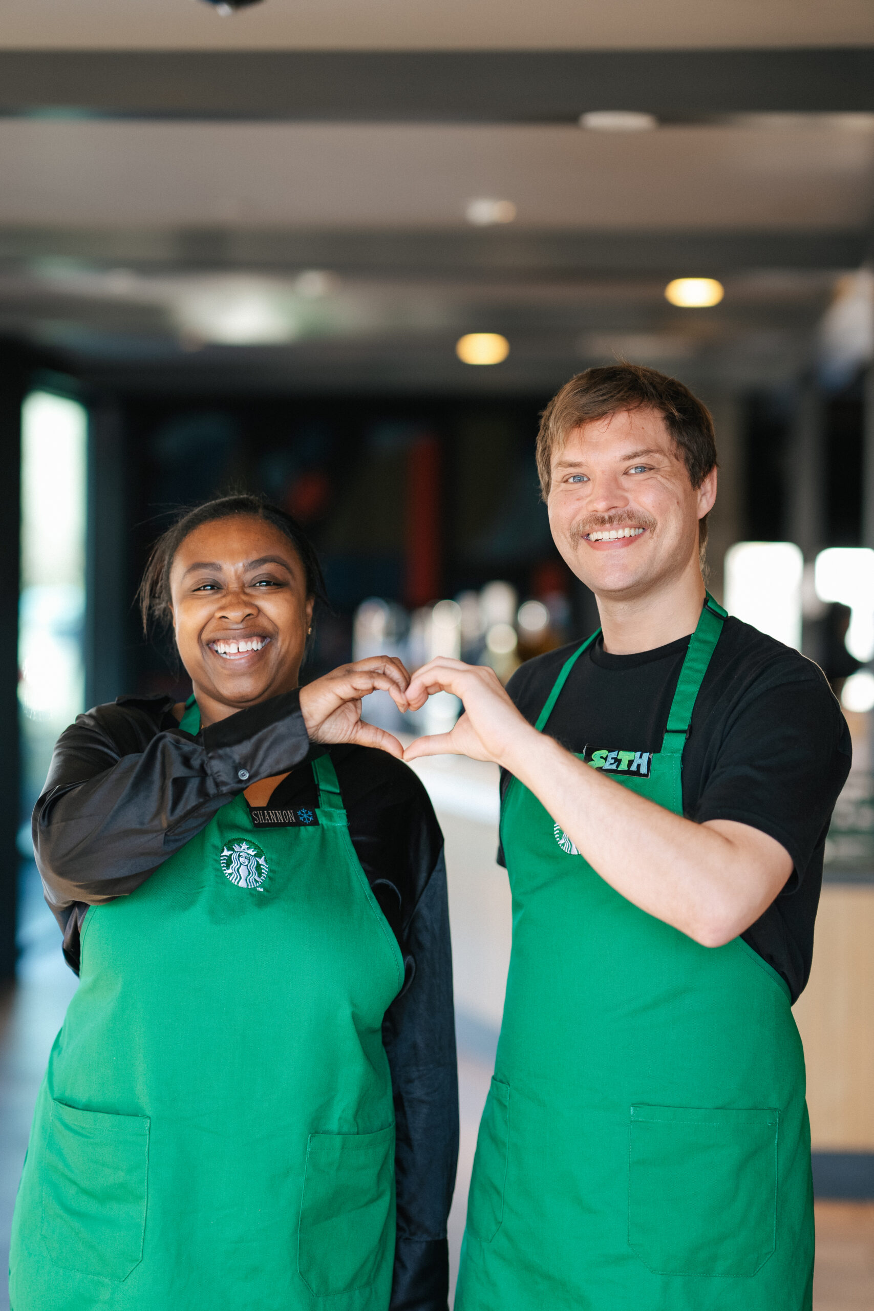 Two baristas are standing together, both wearing green aprons with the Starbucks logo. One is making a heart shape with their hands, while the other is smiling widely. The background features a blurred café setting with warm lighting.