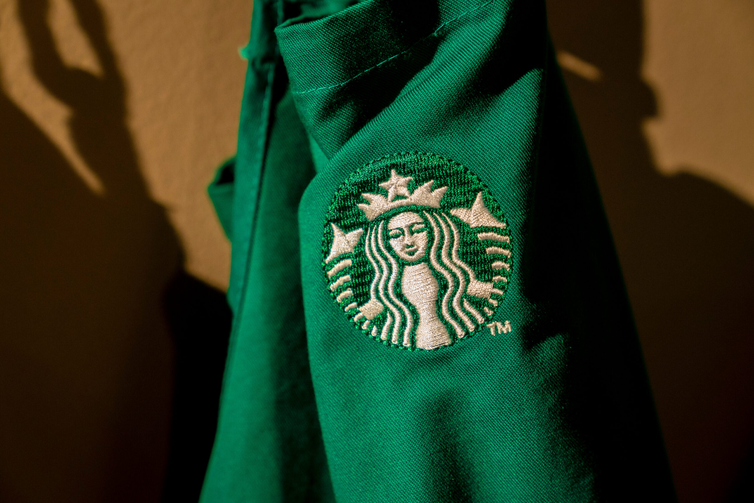 A close-up of a green fabric with an embroidered logo featuring a mermaid-like figure, which is the Starbucks logo. The logo is circular and includes a crown above the figure's head, with wavy hair and a star above. The fabric appears to be part of a garment, likely a uniform, with the logo prominently displayed. The background is a neutral tone, enhancing the focus on the logo.