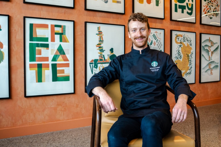 A smiling chef in a black culinary jacket sits in a chair against a backdrop of colorful framed artwork.