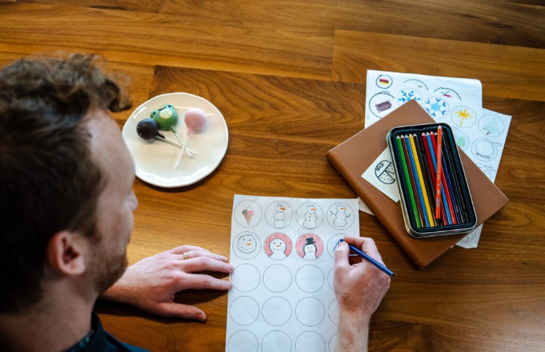 A person is coloring a sheet with snowman illustrations, while a plate with colorful cake pops sits nearby. Colored pencils and a notebook are also on the table.