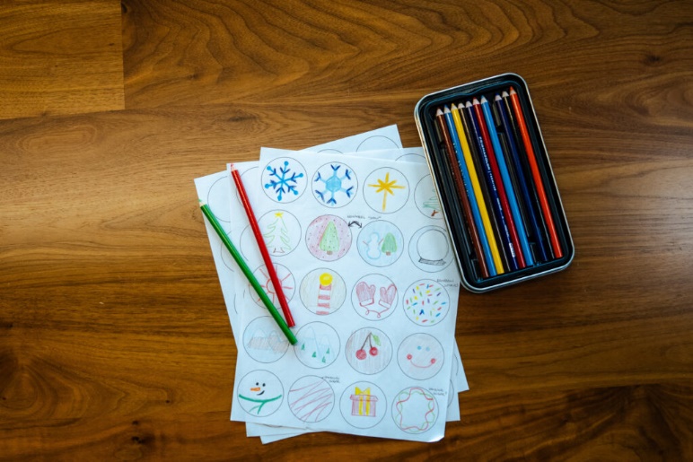 Colored pencils are arranged in a tin next to sheets of paper featuring circular drawings of holiday-themed designs, including snowflakes and trees. Two pencils, one red and one green, rest on the papers. The background is a wooden table.