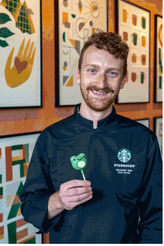 A smiling man in a black chef's jacket holds a green frog-shaped lollipop. Colorful abstract art is displayed on the wall behind him.